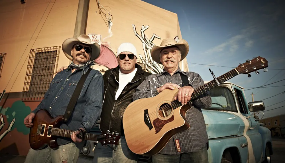 Three musicians stand confidently in front of a vintage blue pickup truck holding their guitars exuding a classic Americana vibe