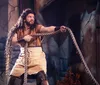 A person is in a theatrical costume holding a thick rope while striking a determined pose on what appears to be a stage set resembling a rustic environment