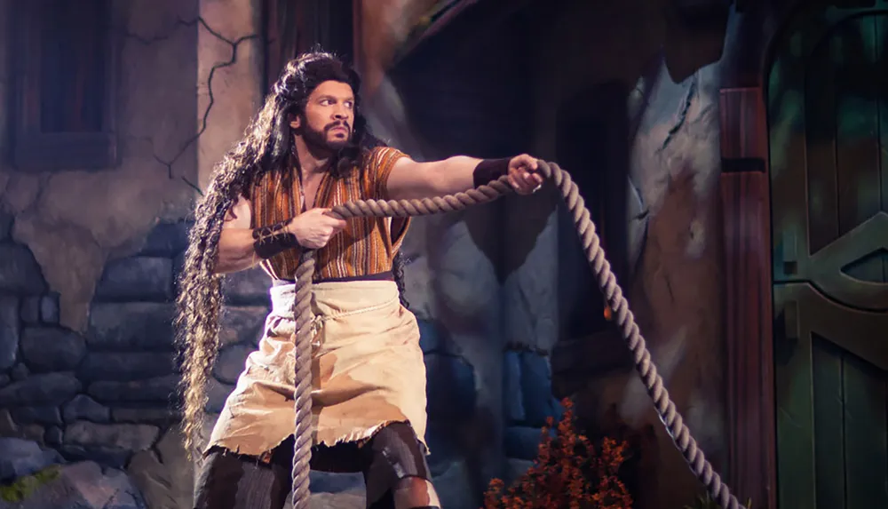 A person is in a theatrical costume holding a thick rope while striking a determined pose on what appears to be a stage set resembling a rustic environment
