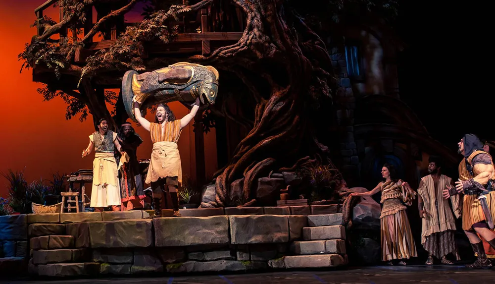 Actors in historical or fantasy costumes are performing on a stage set to resemble an ancient outdoor scene complete with a large tree and stone structures while one actor triumphantly lifts a decorated artifact overhead