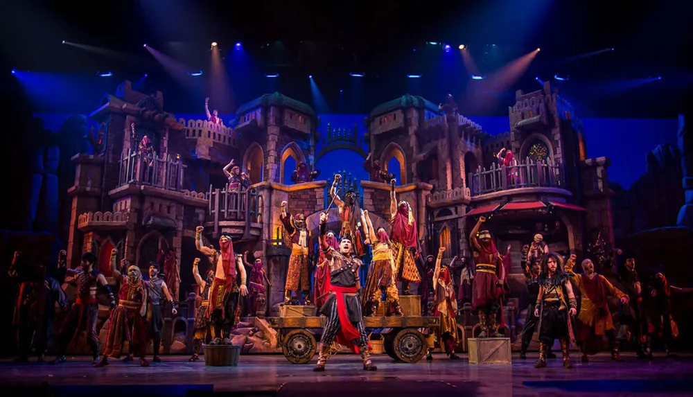 This image depicts a vibrant theatrical production with performers in elaborate costumes on a set designed to resemble a Middle Eastern town likely during a musical number given the dynamic poses and theatrical lighting