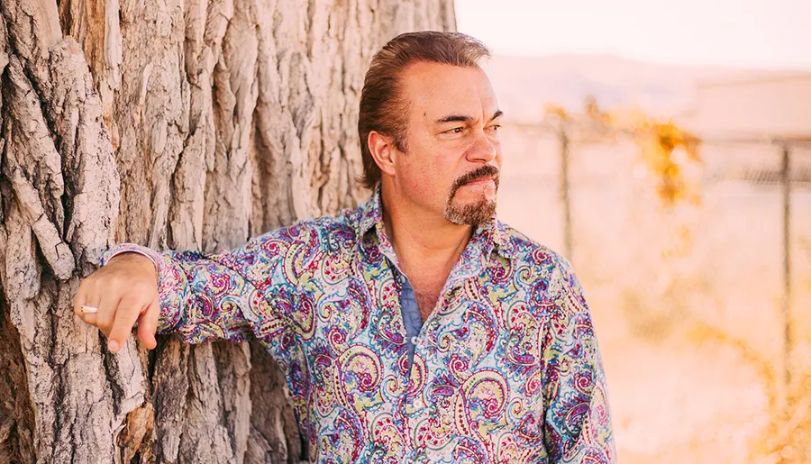 A man with a mustache, wearing a colorful paisley shirt, leans against a tree, gazing pensively into the distance.