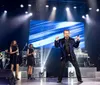 A performer in a blue embellished jacket sings on stage with a band and dancers with the text Bransons Most Popular Morning Show