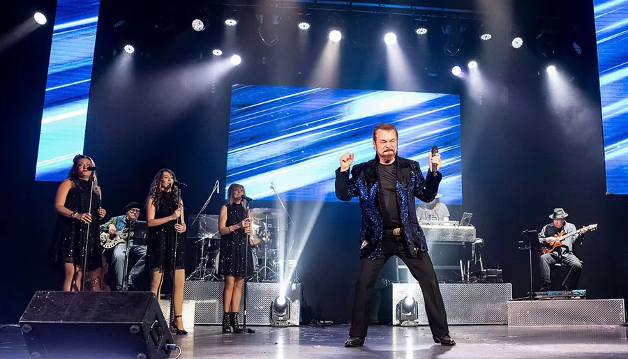 A performer energetically sings on stage with a live band and backup singers, illuminated by spotlights and blue screen visuals.