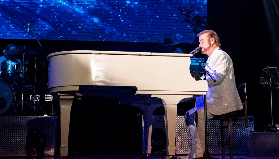 A man in a white suit is playing a white grand piano on stage, singing into a microphone, with a band and blue-lit background.
