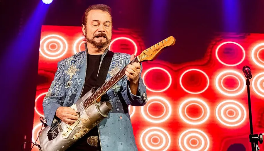 A musician in a decorated jacket is playing an electric guitar on stage with a vibrant red and orange circular light pattern in the background.