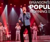 A performer in a blue embellished jacket sings on stage with a band and dancers with the text Bransons Most Popular Morning Show