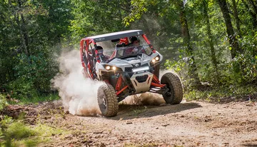 A rugged off-road vehicle kicks up dust as it speeds along a dirt trail through a forested area.