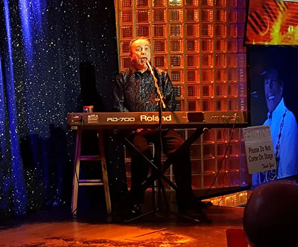 A musician is performing on stage singing into a microphone while playing a Roland RD-700 keyboard set against a backdrop of sparkly blue curtains and decorative panels