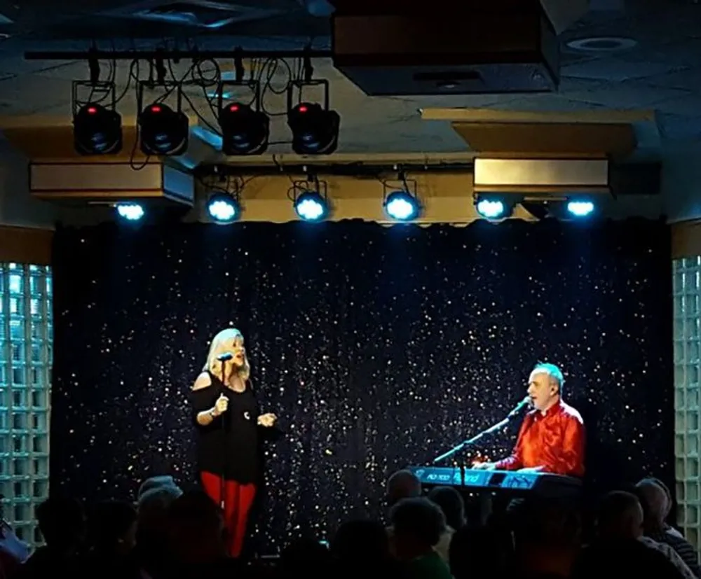 A singer performs next to a pianist on a small stage adorned with blue lights and a starry backdrop in front of an audience