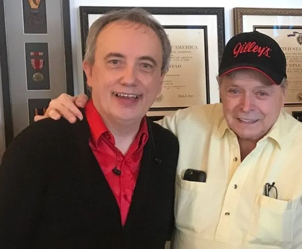 Two smiling men are posing for a photo with one wearing a black jacket over a red shirt and the other in a cream-colored shirt and a black cap with Gilleys written on it