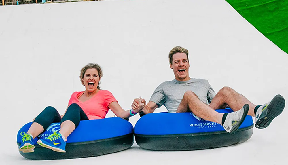 Two people are joyfully sliding down a synthetic slope on inner tubes at an outdoor recreation area