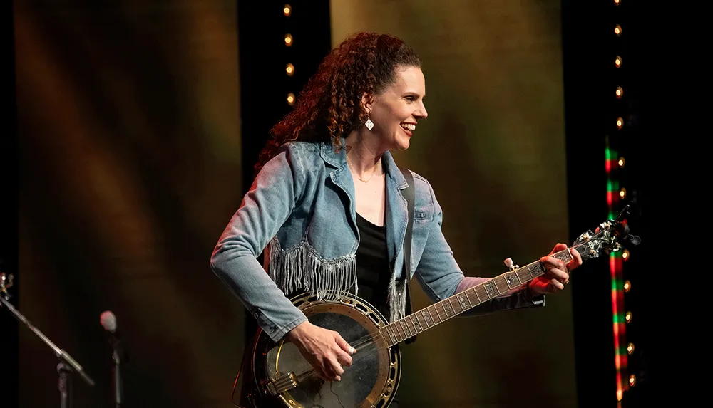 A person with curly hair is smiling and playing a banjo on stage
