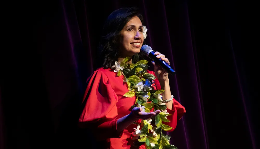 A woman wearing a red dress and a floral lei is speaking into a microphone on stage