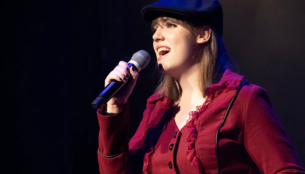 A person wearing a black hat and a maroon coat is singing into a microphone on stage