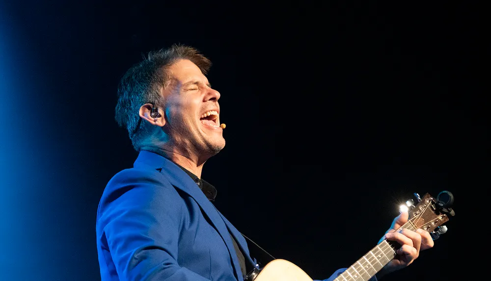 A person in a blue blazer is passionately singing while playing an acoustic guitar on stage