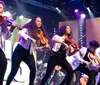 A group of musicians energetically plays violins on stage wearing matching white jackets with colorful lights in the background