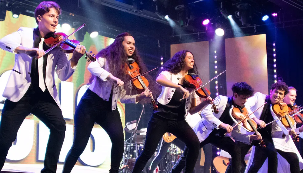 A group of musicians energetically plays violins on stage wearing matching white jackets with colorful lights in the background