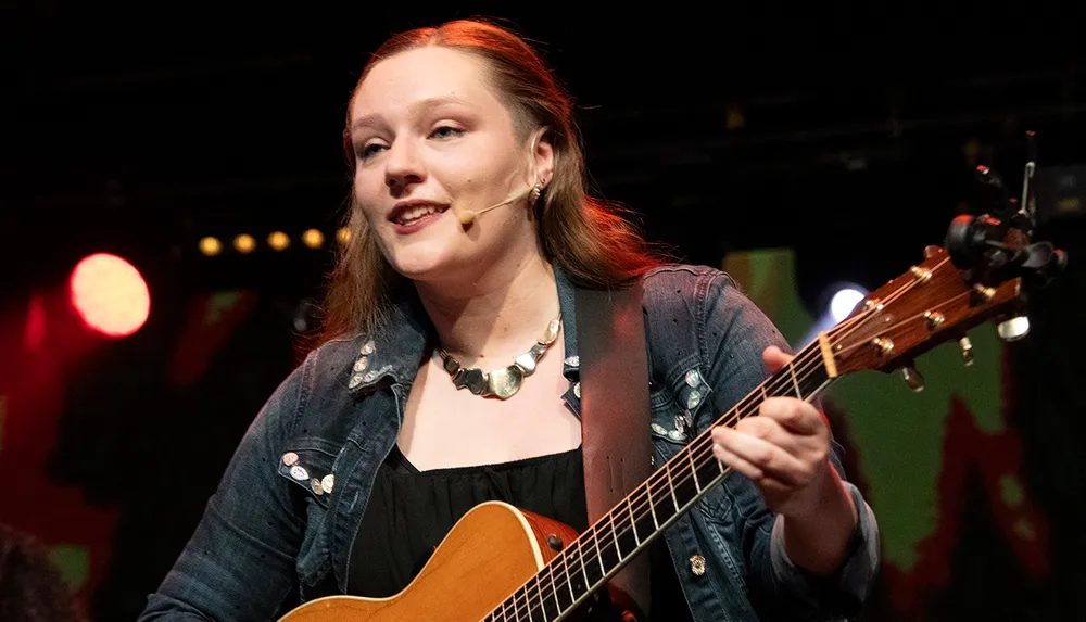 A person is playing guitar on stage with a microphone headset and warm stage lighting in the background