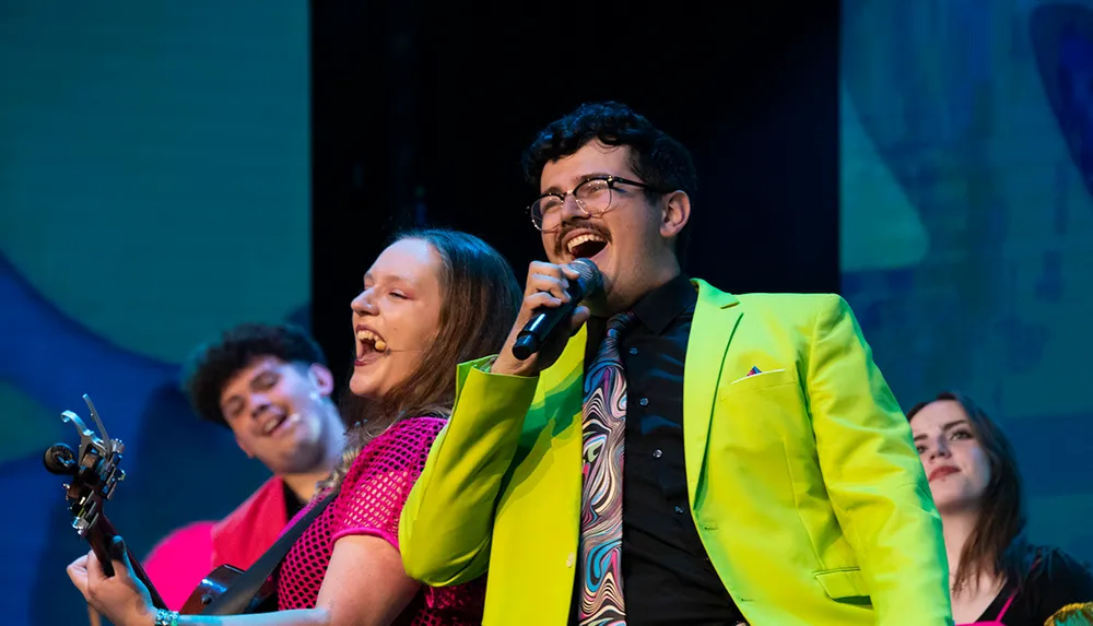 A vibrant group of performers energetically sing and play on stage with one wearing a bright neon suit and another playing guitar