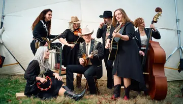 A group of musicians wearing Western-style outfits is performing with various instruments, including guitars, a violin, a mandolin, and a double bass, in a casual outdoor setting.