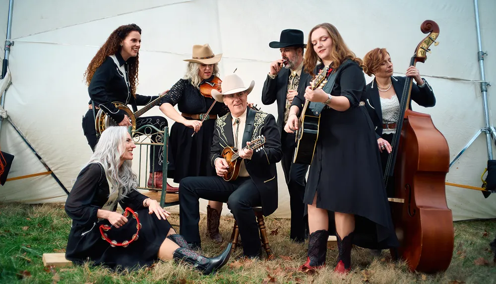A group of musicians wearing Western-style outfits is performing with various instruments including guitars a violin a mandolin and a double bass in a casual outdoor setting