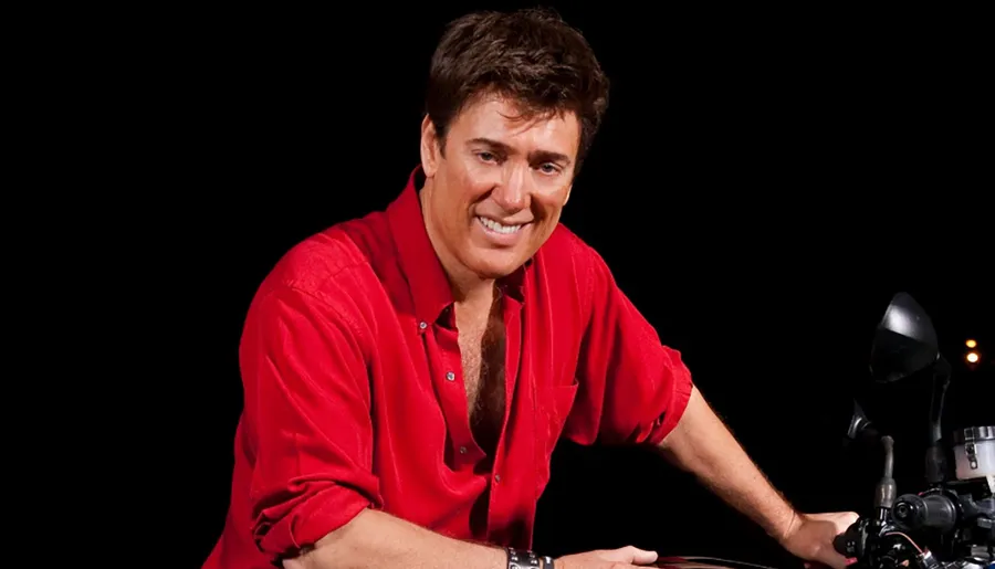 A person with a cheerful smile is dressed in a red shirt and seated on a motorcycle, set against a dark background.