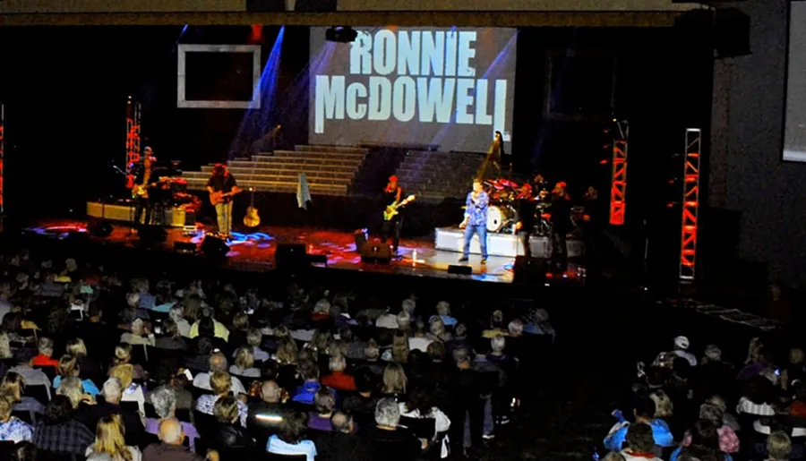 A live concert is taking place in front of an audience with the name Ronnie McDowell projected in large letters on the screen behind the stage.