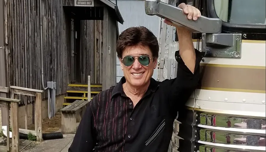 A person with a cheerful expression is standing next to a vehicle, wearing sunglasses and holding onto a part of the vehicle, with a rustic wooden building in the background.