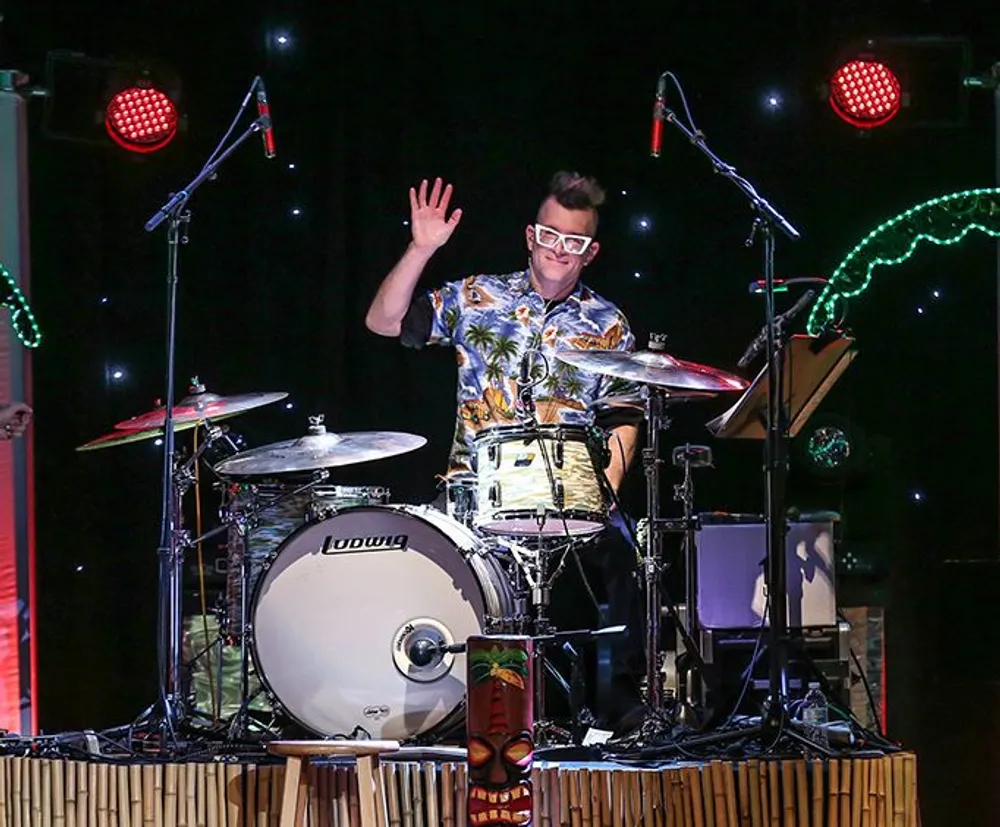 A person wearing sunglasses and a Hawaiian shirt is seated behind a drum set waving with an upbeat expression