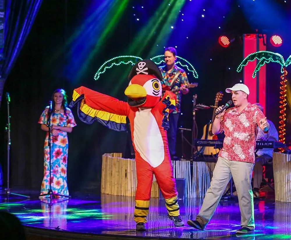 A person in a colorful parrot costume with a pirate hat dances on a stage next to a man singing into a microphone with other performers in tropical attire and illuminated palm trees in the background