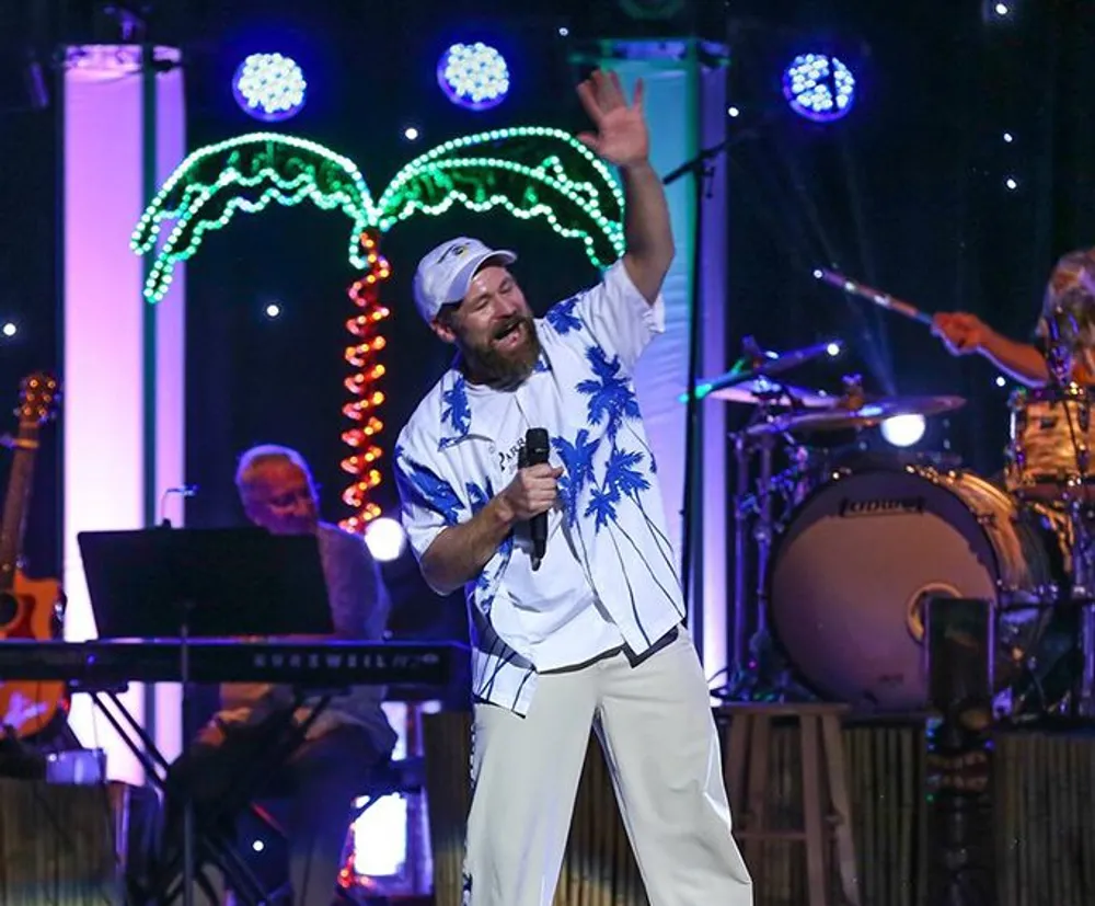 A man in a tropical-themed shirt and white pants is enthusiastically singing or speaking into a microphone on stage with musicians in the background