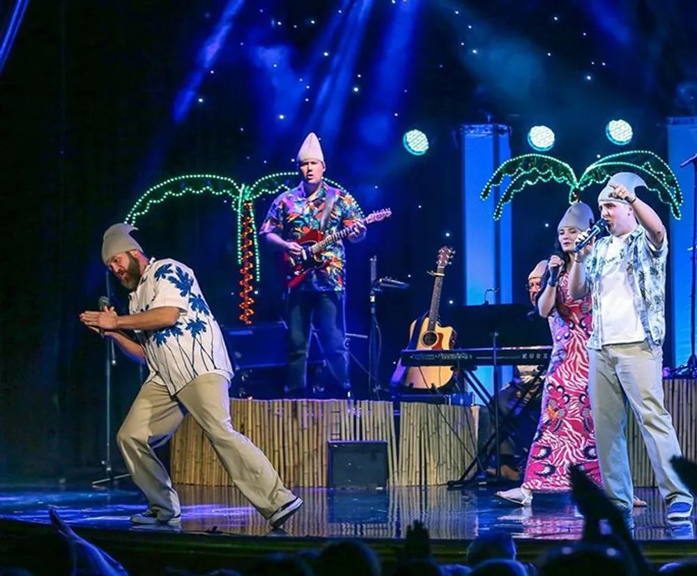 Performers on stage engage in a lively act with a tropical theme while one plays the guitar and two others dance in colorful outfits