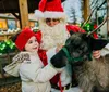 A child in winter clothing smiles while touching a reindeer as an individual dressed as Santa Claus stands beside them in a festive setting