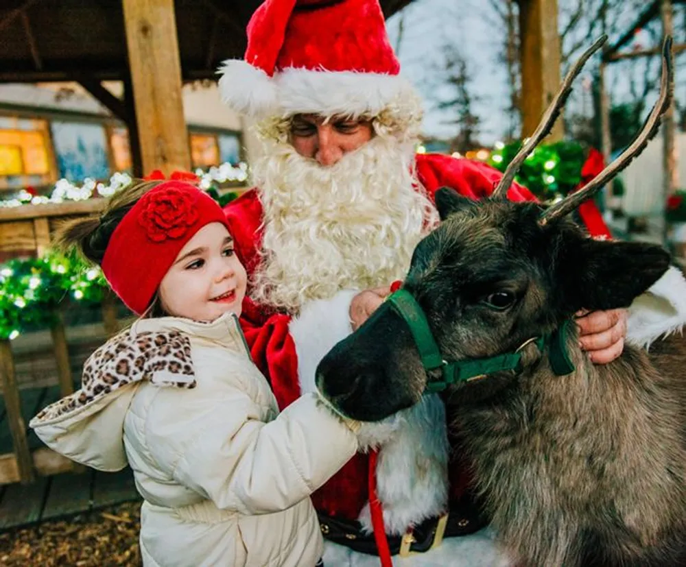 A child in winter clothing smiles while touching a reindeer as an individual dressed as Santa Claus stands beside them in a festive setting