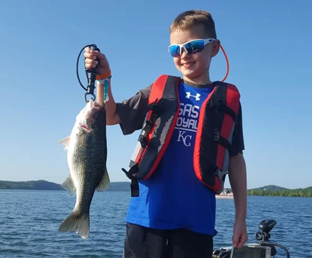 A boy wearing sunglasses and a life vest proudly displays a fish he has caught with a lake and clear sky in the background