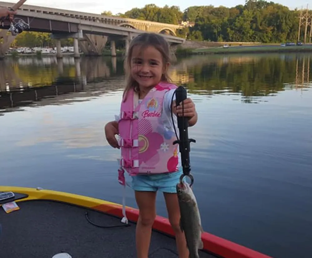 A smiling young girl wearing a pink life jacket is proudly holding up a fish she caught while standing on a boat