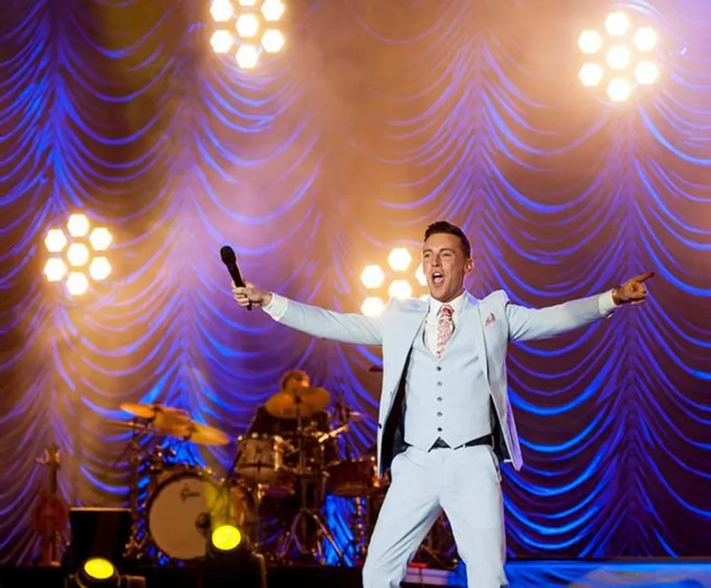 A performer in a white suit energetically sings on stage with outstretched arms backed by theatrical purple lighting and a drummer in the background