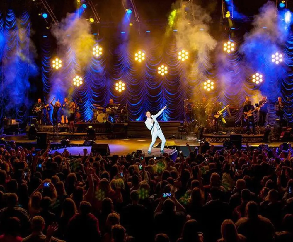 A performer is singing on stage in front of a large audience with vivid stage lighting and a live band in the background