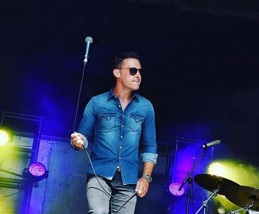The image shows a man on stage wearing sunglasses and holding a microphone in one hand appearing to be performing or preparing to perform with musical equipment in the background