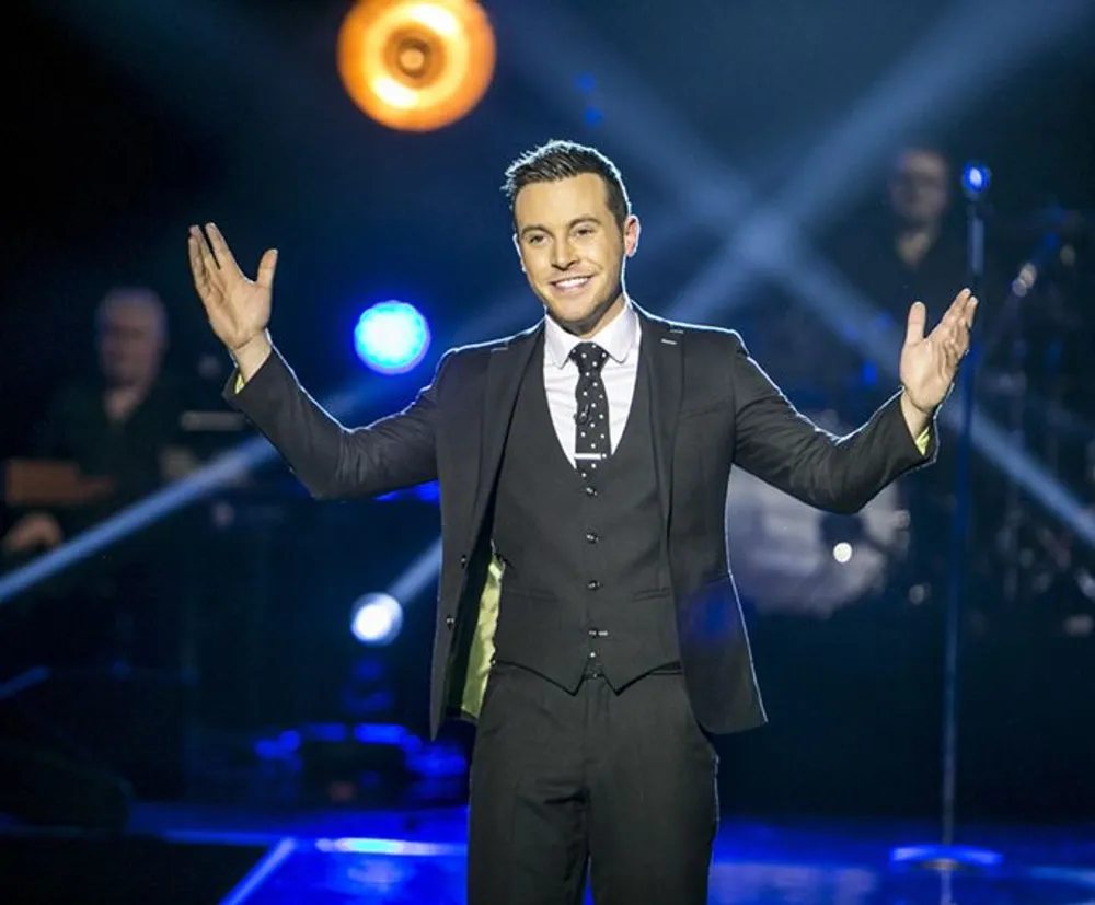 A person in a suit is standing on stage with their arms open wide and smiling with stage lights and musicians in the background