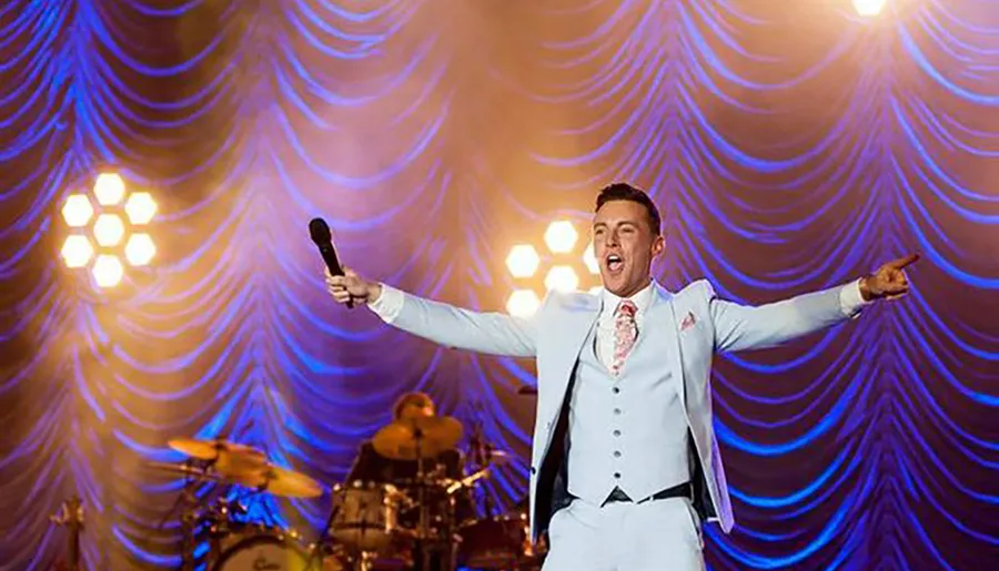 A performer in a white suit with arms outstretched sings into a microphone on a stage with purple lighting and a band in the background.