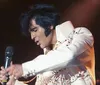 A performer in a gold jacket passionately sings into a vintage microphone on stage with a guitar slung across their shoulder