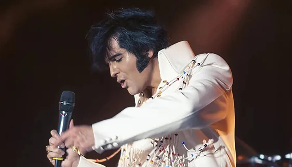 A performer in a white fringed outfit passionately sings into a microphone under stage lights