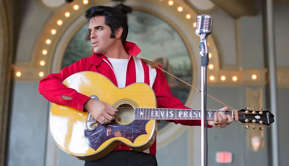 A person dressed in a red and white jacket is posing with an acoustic guitar in front of a vintage microphone set against a decorative backdrop