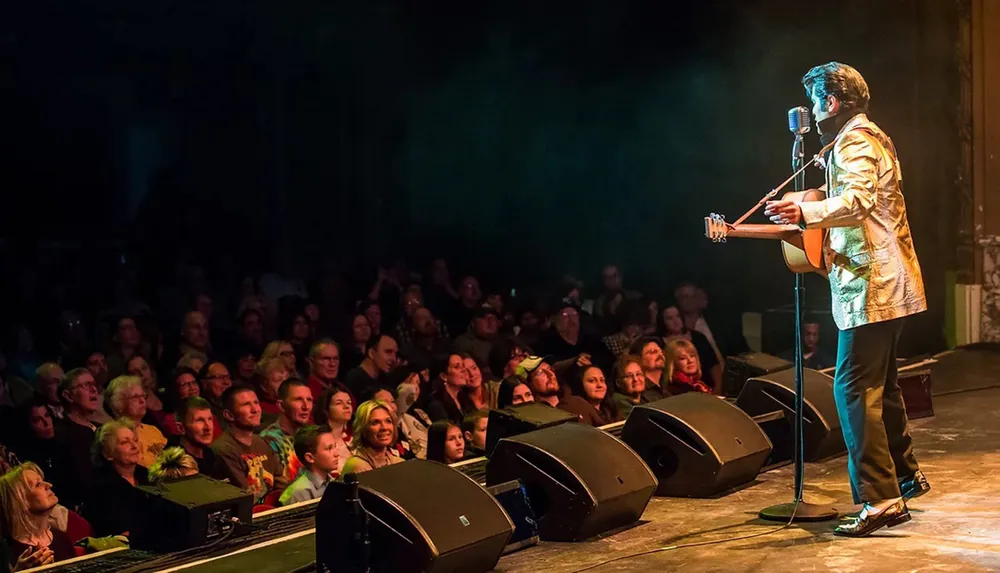 A performer in a gold jacket plays the guitar and sings into a microphone on stage engaging an attentive audience