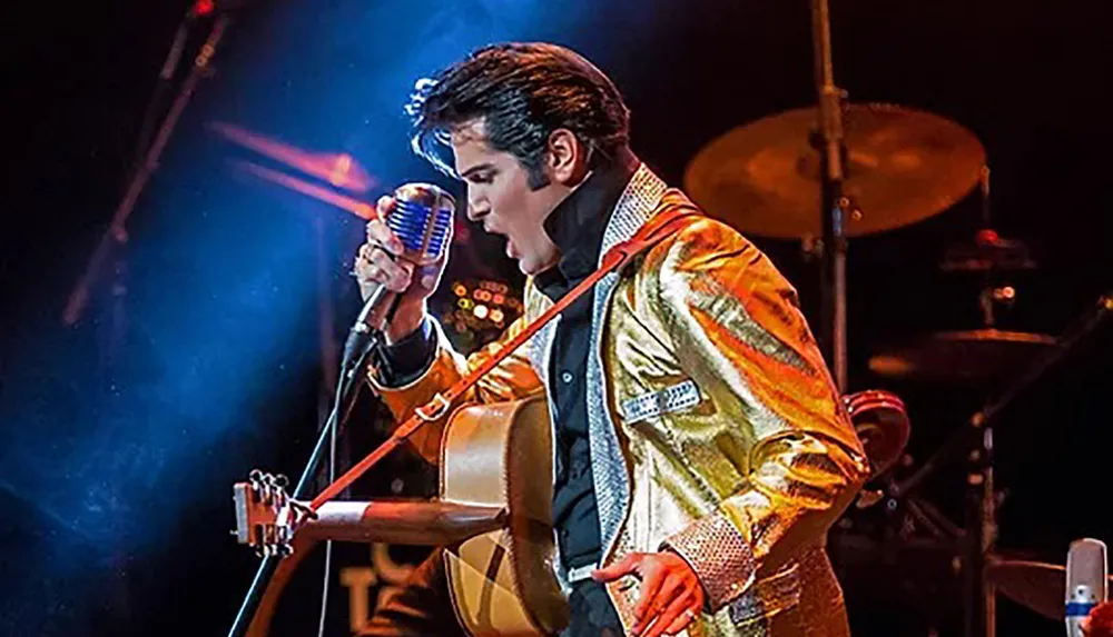 A performer in a gold jacket passionately sings into a vintage microphone on stage with a guitar slung across their shoulder