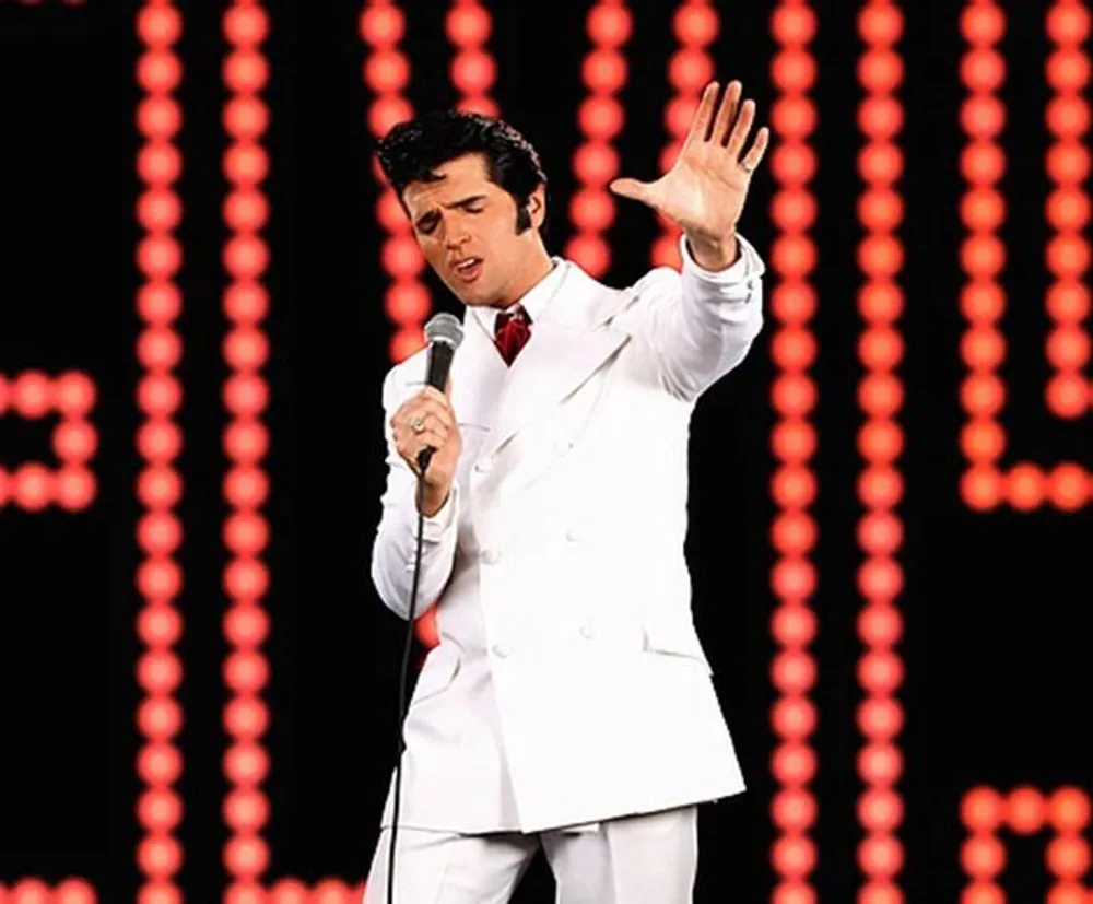 A performer dressed in a white suit and red tie is singing into a microphone with his left hand raised against a background with red light patterns