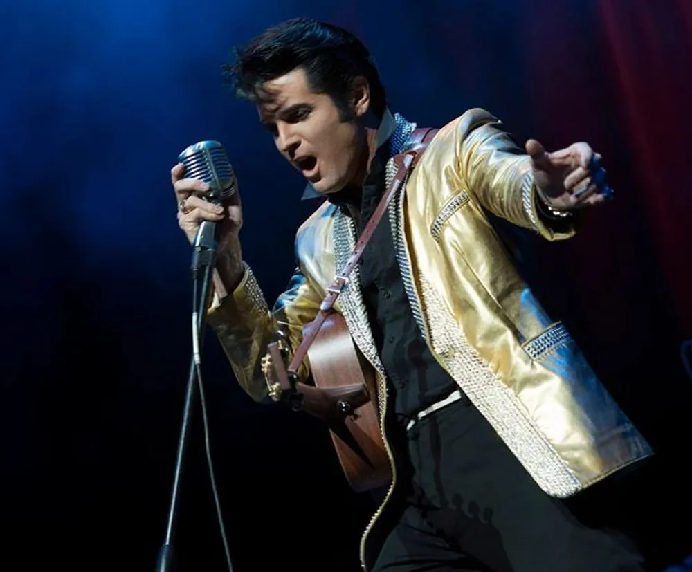 A person dressed in an elaborate gold and black outfit performs with a guitar and a vintage microphone exuding a strong stage presence reminiscent of classic rock and roll