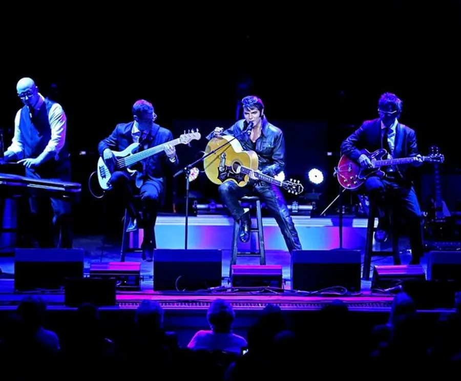A band consisting of four musicians performs on stage with a focus on the central figure playing an acoustic guitar.